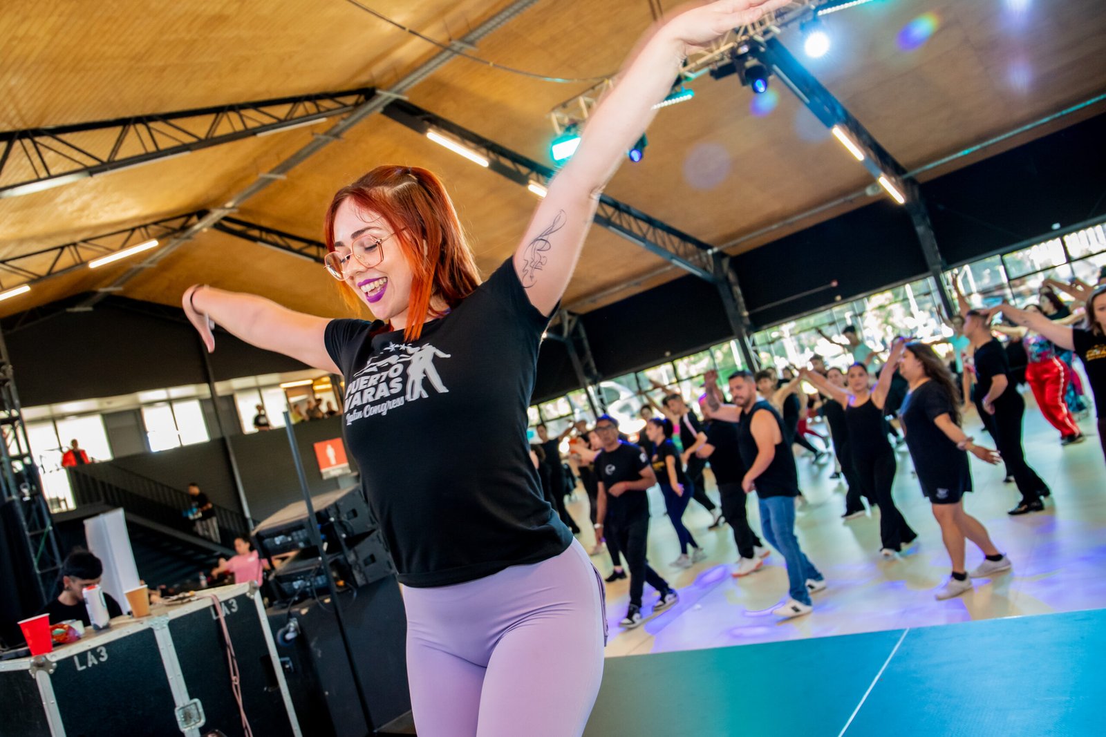 Irene Palomares teaching a workshop: smiling with her arms open as she calls the step, while dozens of students dance behind her